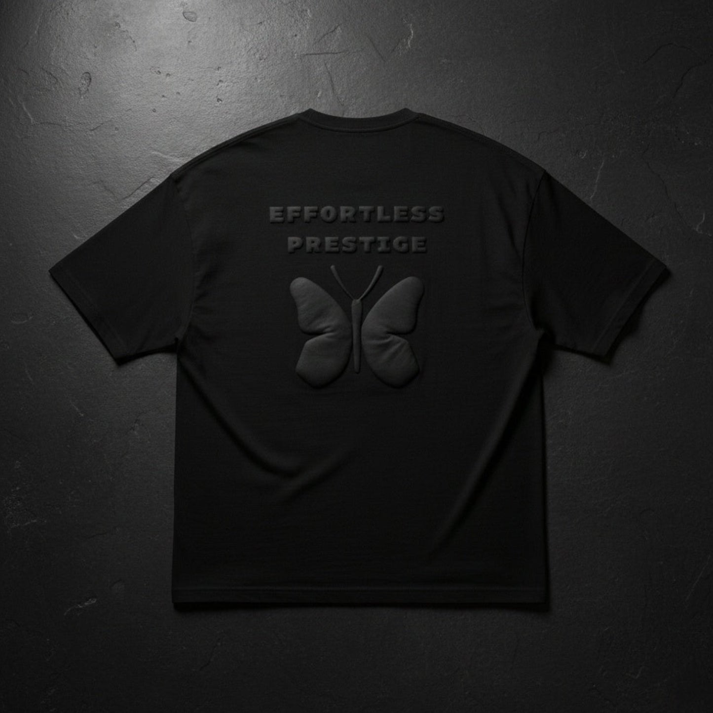 Black oversized t-shirt laid flat with embossed butterfly motif and ‘Effortless Prestige’ text, dark textured background.
