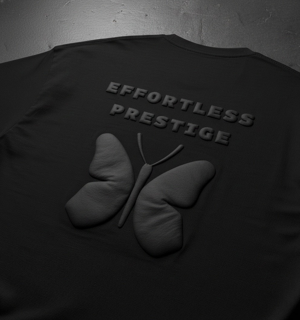 Black oversized t-shirt close-up showing embossed butterfly graphic and ‘Effortless Prestige’ text, textured lighting on dark surface.