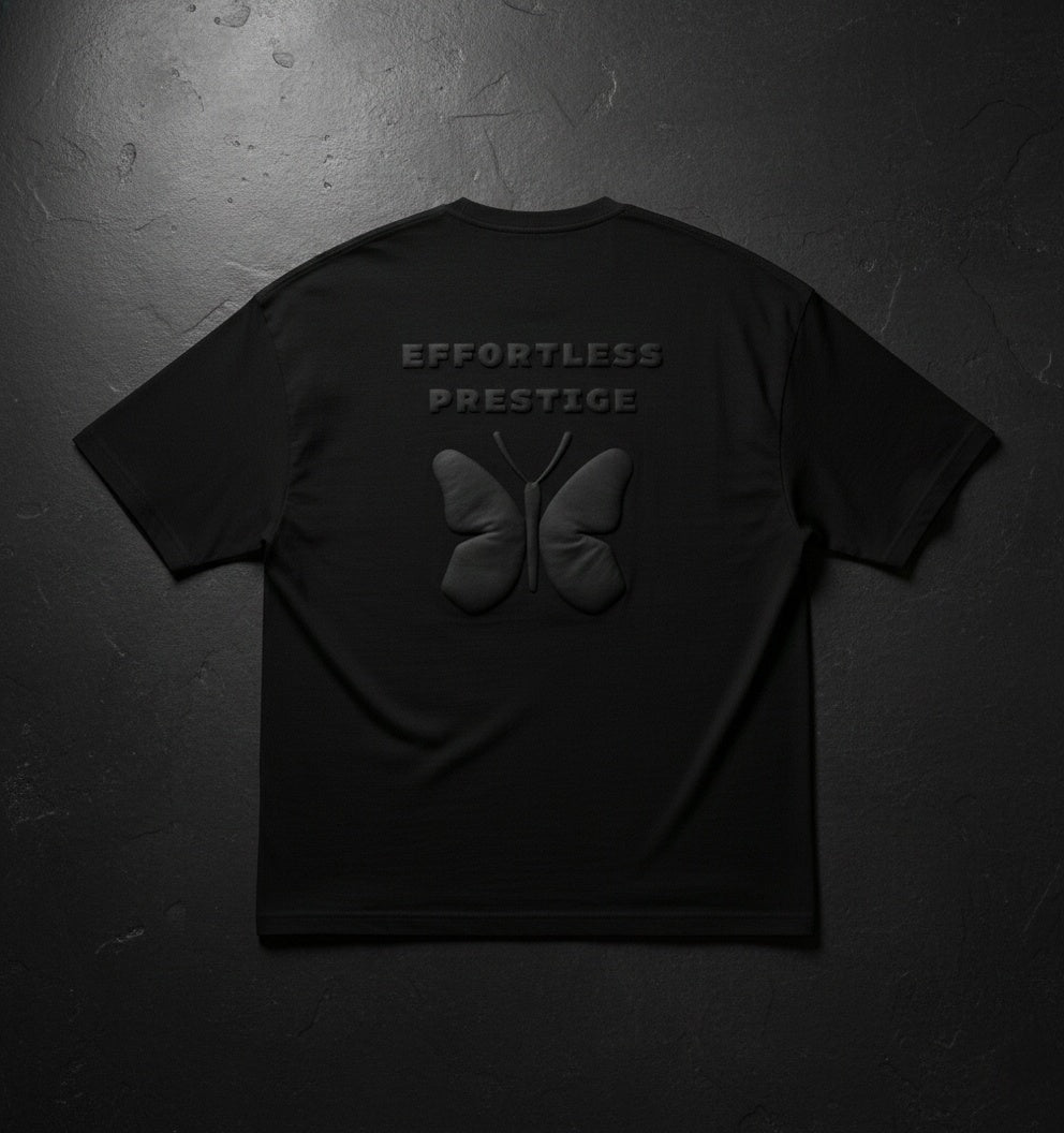 Black oversized t-shirt laid flat with embossed butterfly motif and ‘Effortless Prestige’ text, dark textured background.
