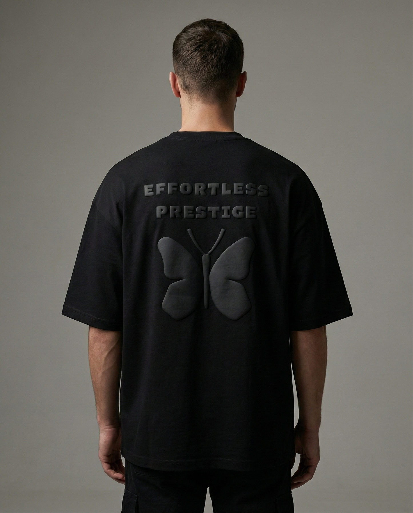 Male model wearing black oversized Effortless Prestige t-shirt with raised butterfly design and embossed lettering, studio background.
