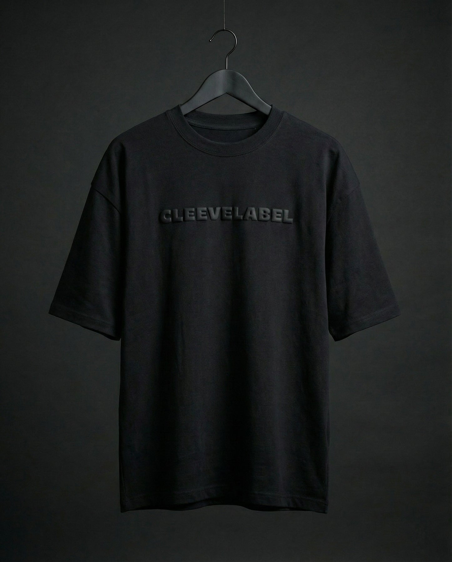 Black oversized t-shirt on hanger with raised Cleevelabel text on front, dark minimal studio backdrop.