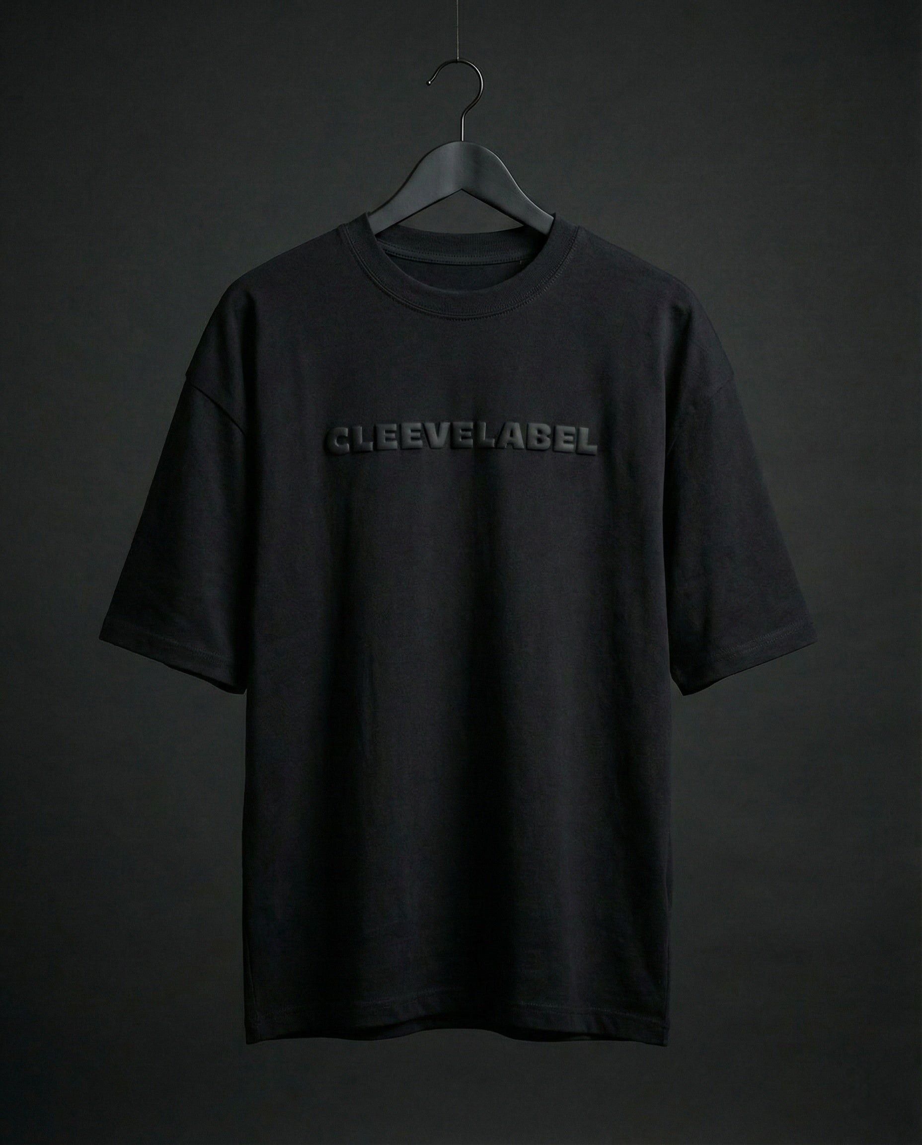 Black oversized t-shirt on hanger with raised Cleevelabel text on front, dark minimal studio backdrop.