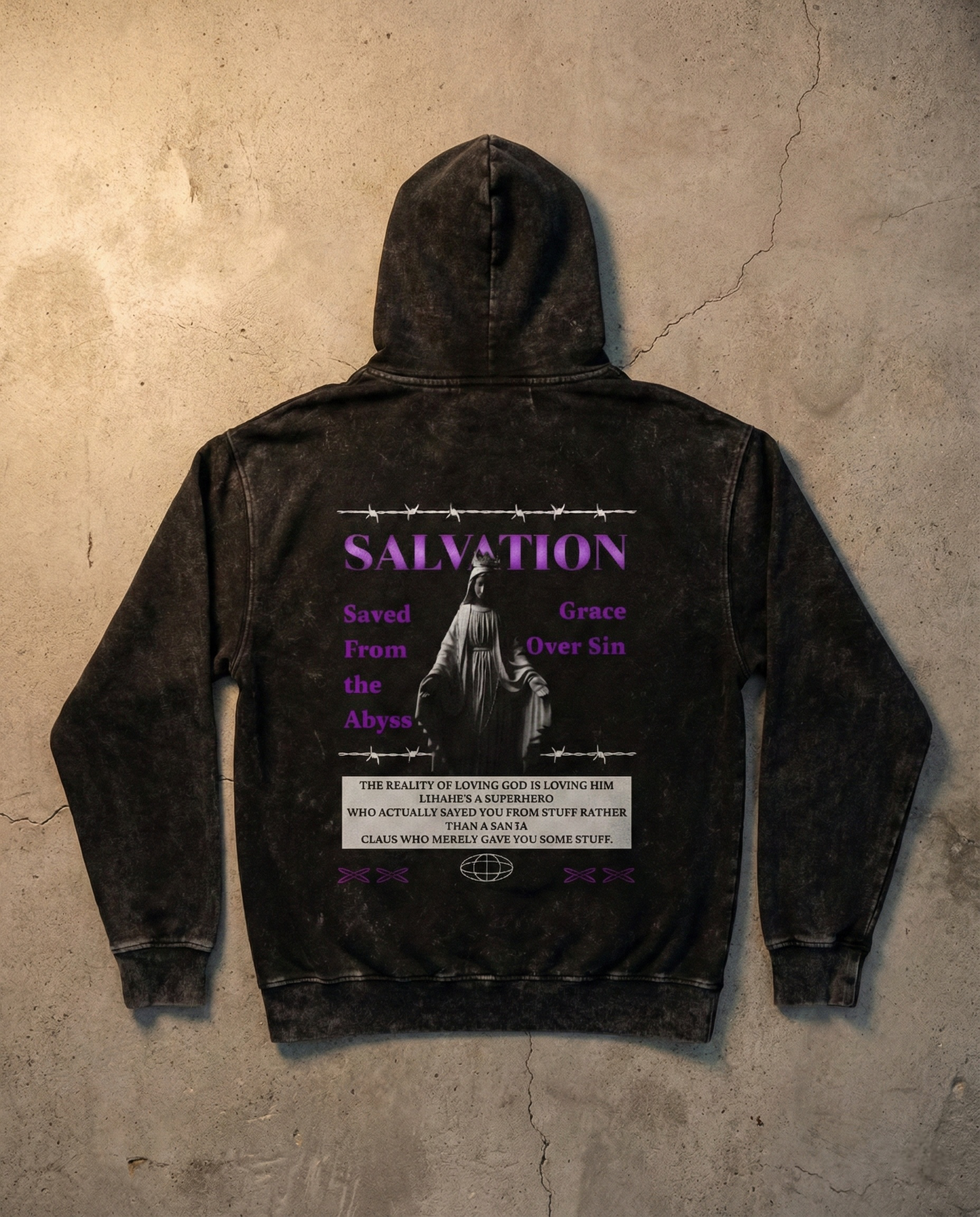 Black acid-wash Salvation hoodie laid flat on concrete surface, back view featuring purple typography, statue artwork, and barbed wire details.